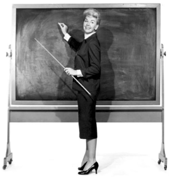 Vintage image in black and white of a white woman with blond hair pulled up wearing a black skirt, jacket, and pumps standing in front of a chalkboard with a long pointing stick in her left hand and looking like she will start writing with chalk that is in her right hand. Her body is perpendicular to the chalkboard and she is looking over her left shoulder at the camera. 