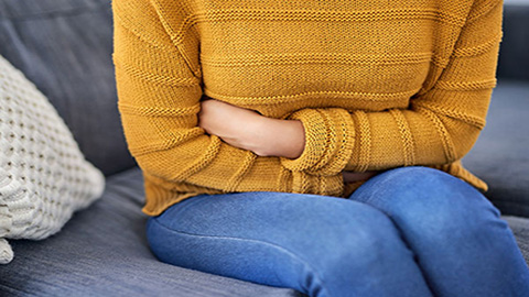 Woman in a burnt orange sweater and jeans sitting on a gray couch with her arms crossed over her abdomen