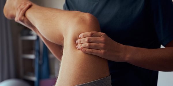 A massage therapist holding a right knee up in the air at a 90 degree angle with the therapist's left hand squeezing just above the knee and their right hand supporting the leg under the calf.